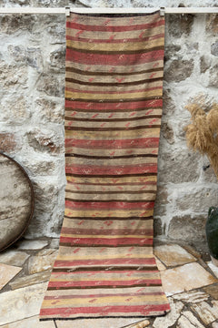 A long striped kilim textile hangs from a white rod against a rustic stone wall, with a dark green ceramic vase of dried pampas grass and a round wooden barrel prop on the floor.